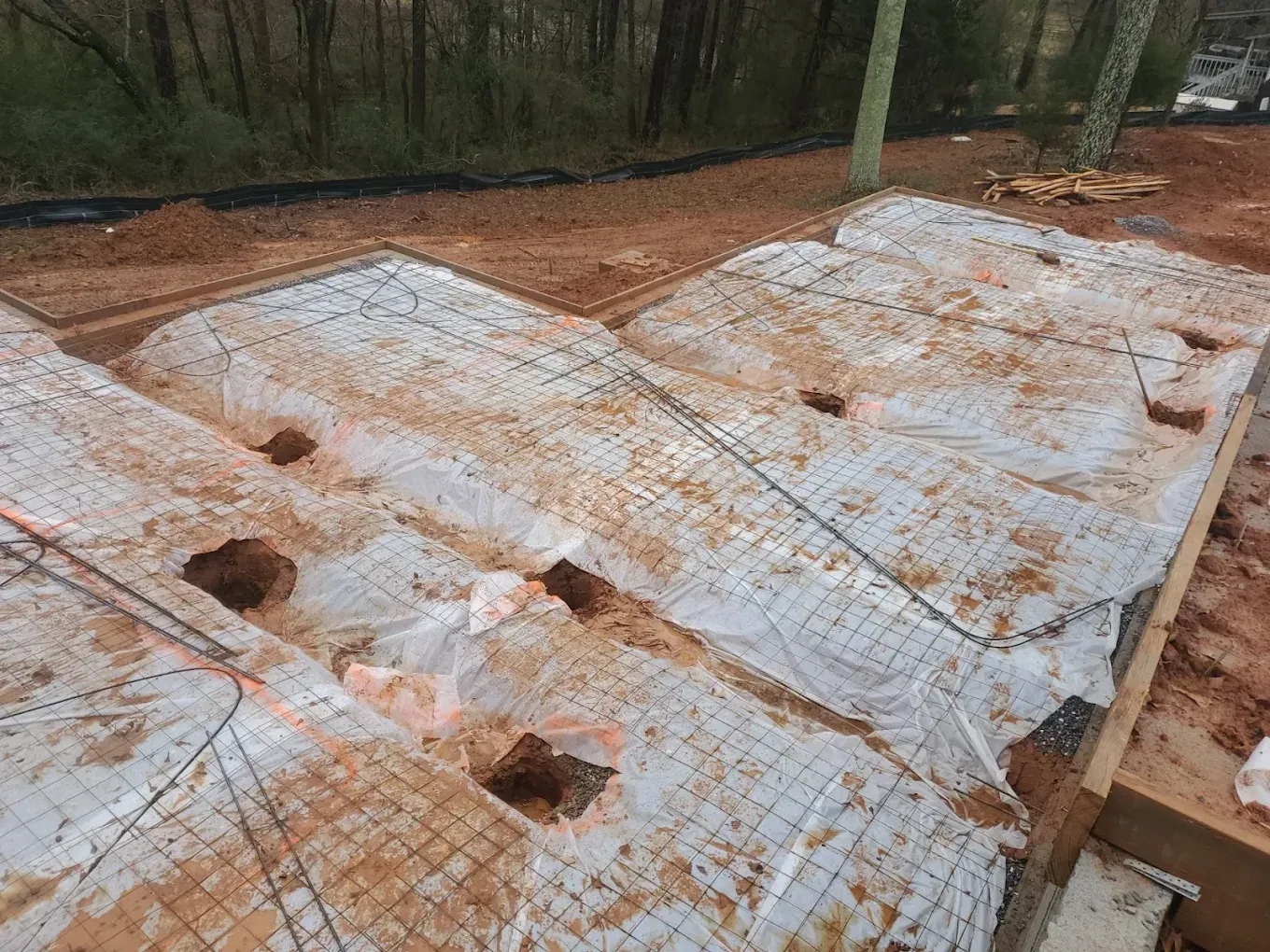 Construction site with wood framing, mesh, and holes; covered by white material, brown dirt, and trees in the background.