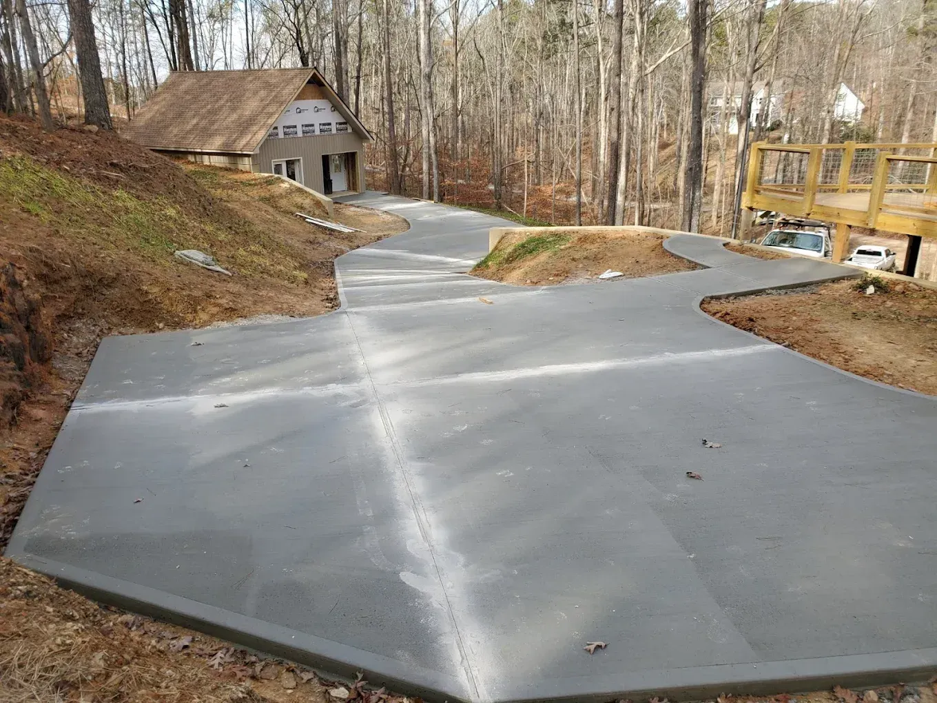 A gray concrete driveway curves up a hill toward a house under construction, surrounded by trees.