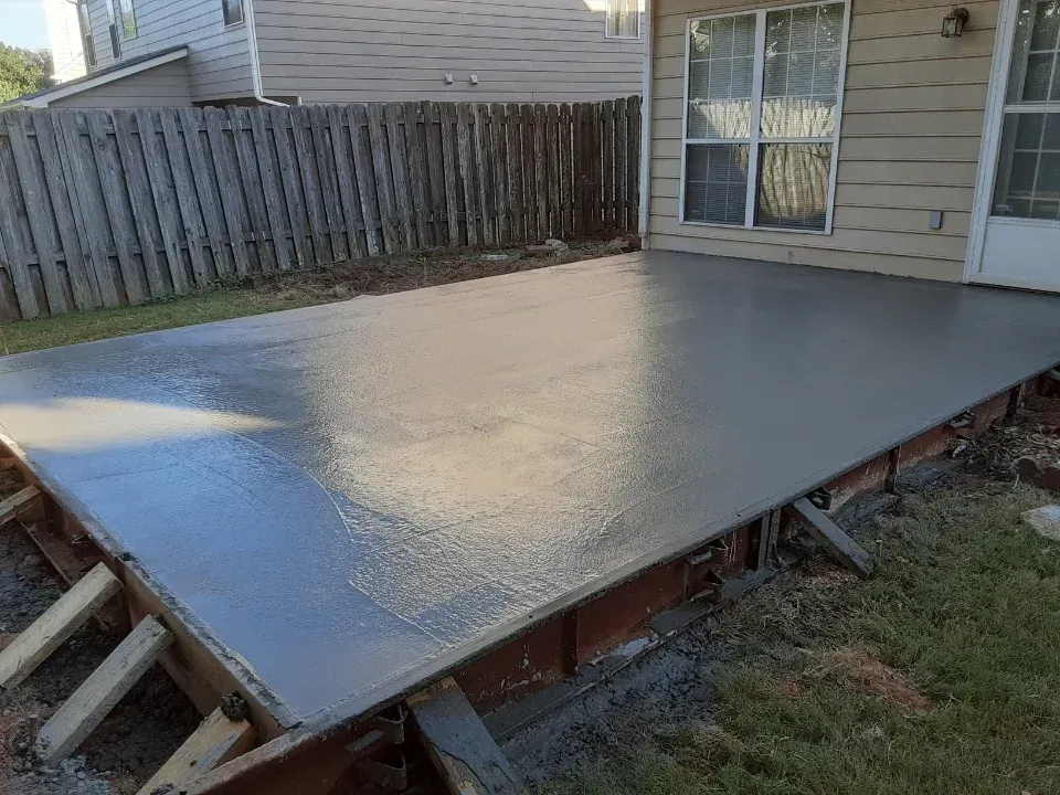 Newly poured concrete patio with wooden framework, next to a house and fence.