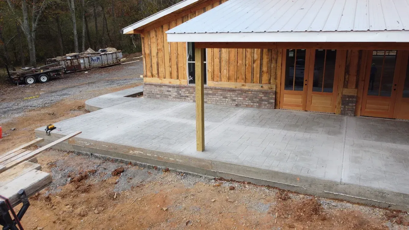 Concrete patio being poured in front of a log cabin with wood doors, and a trailer of supplies nearby.