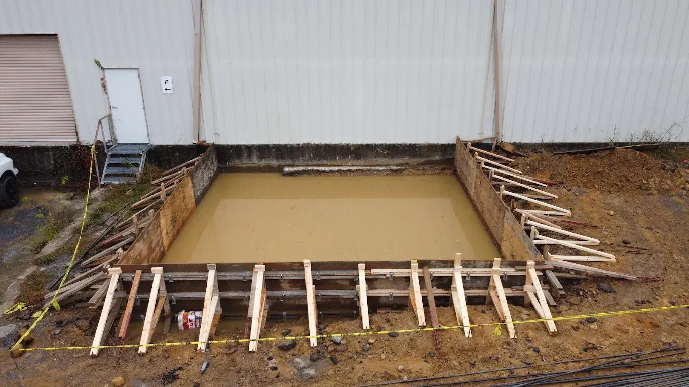 Construction site: Wooden frame surrounding muddy pit in front of a building with a door and steps.
