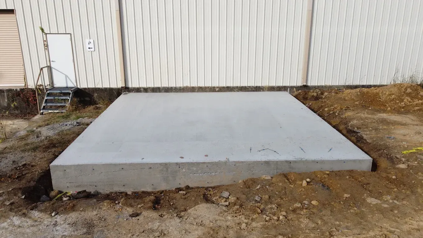 Concrete pad in front of a building with a white door and metal siding, surrounded by dirt.