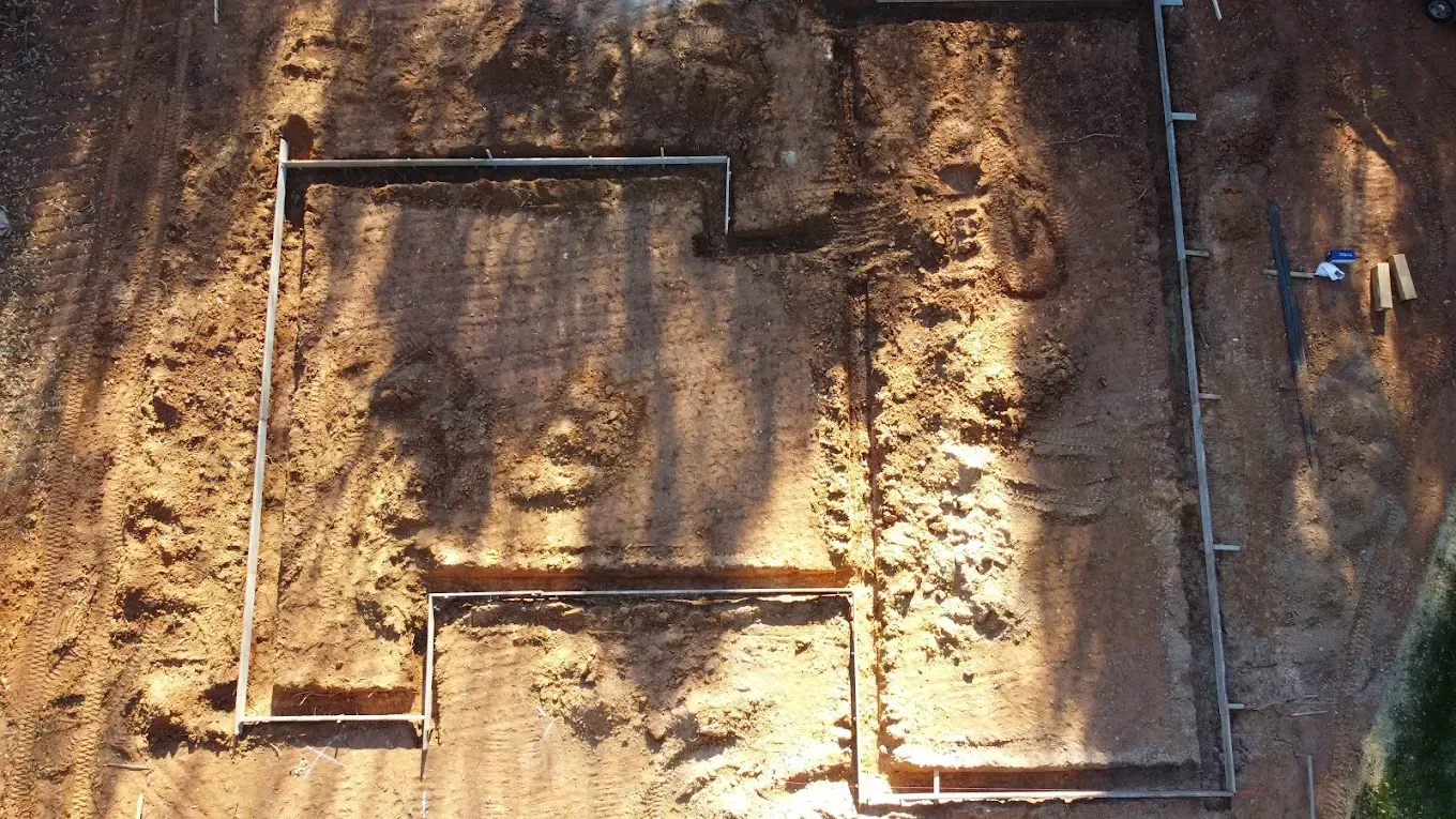 Overhead view of a construction site with foundation trenches marked by metal pipes in the dirt.