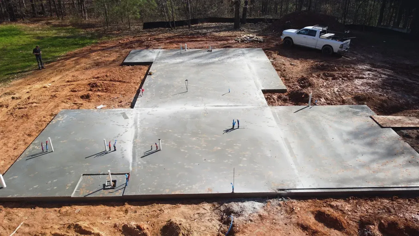 Concrete foundation poured for a house, with plumbing pipes visible. A truck and person stand nearby.
