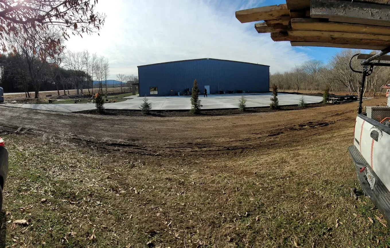 Blue warehouse building on a concrete pad, surrounded by dirt and a row of young trees; daytime.