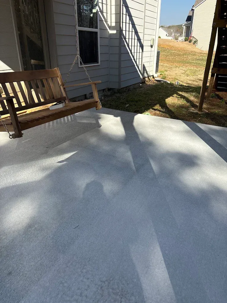 Wooden porch swing near a house, with shadows on a concrete patio, in sunlight.