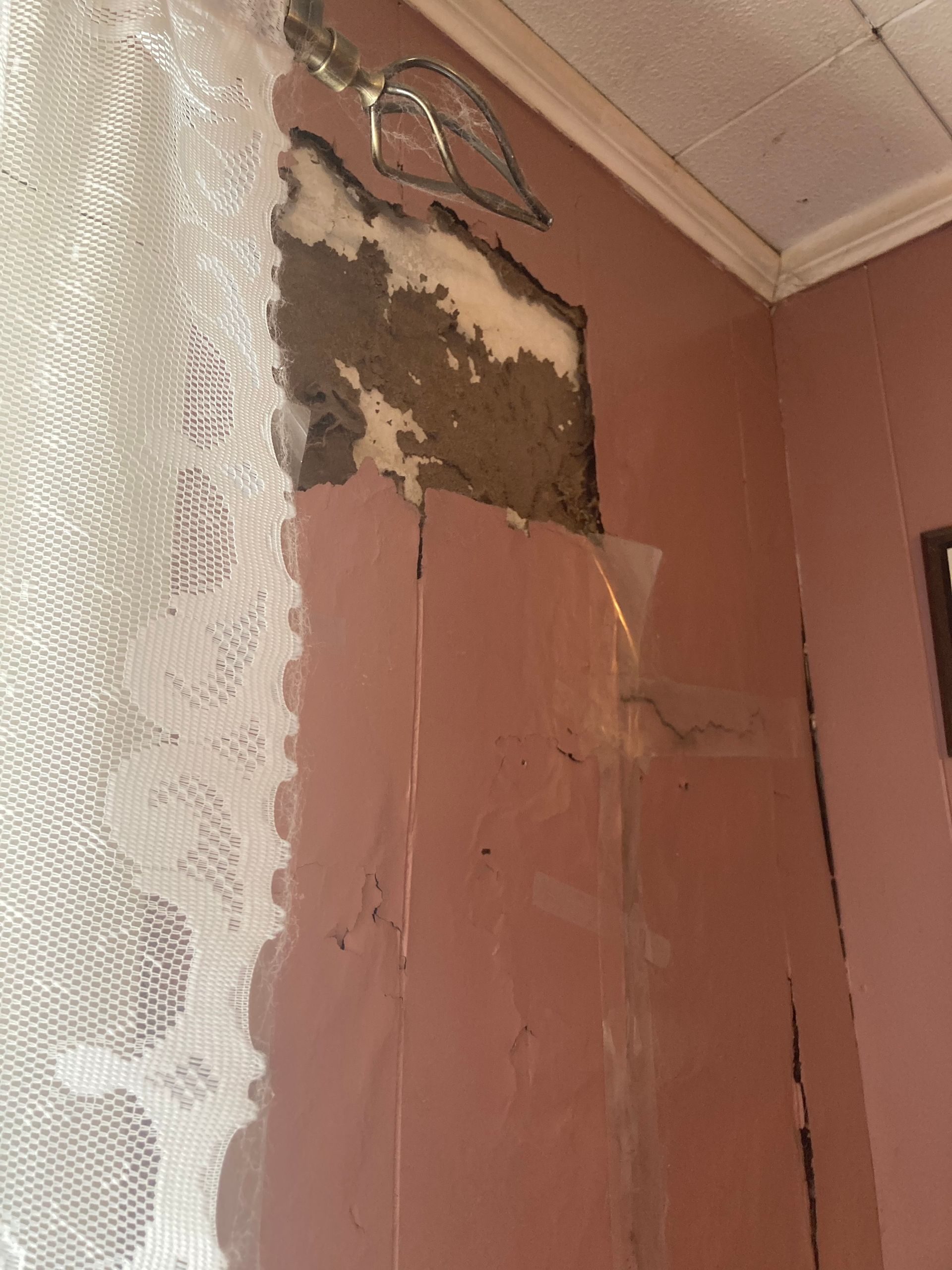 Damaged pink wall with exposed plaster, curtain in the left, white ceiling, and a few electrical wires.