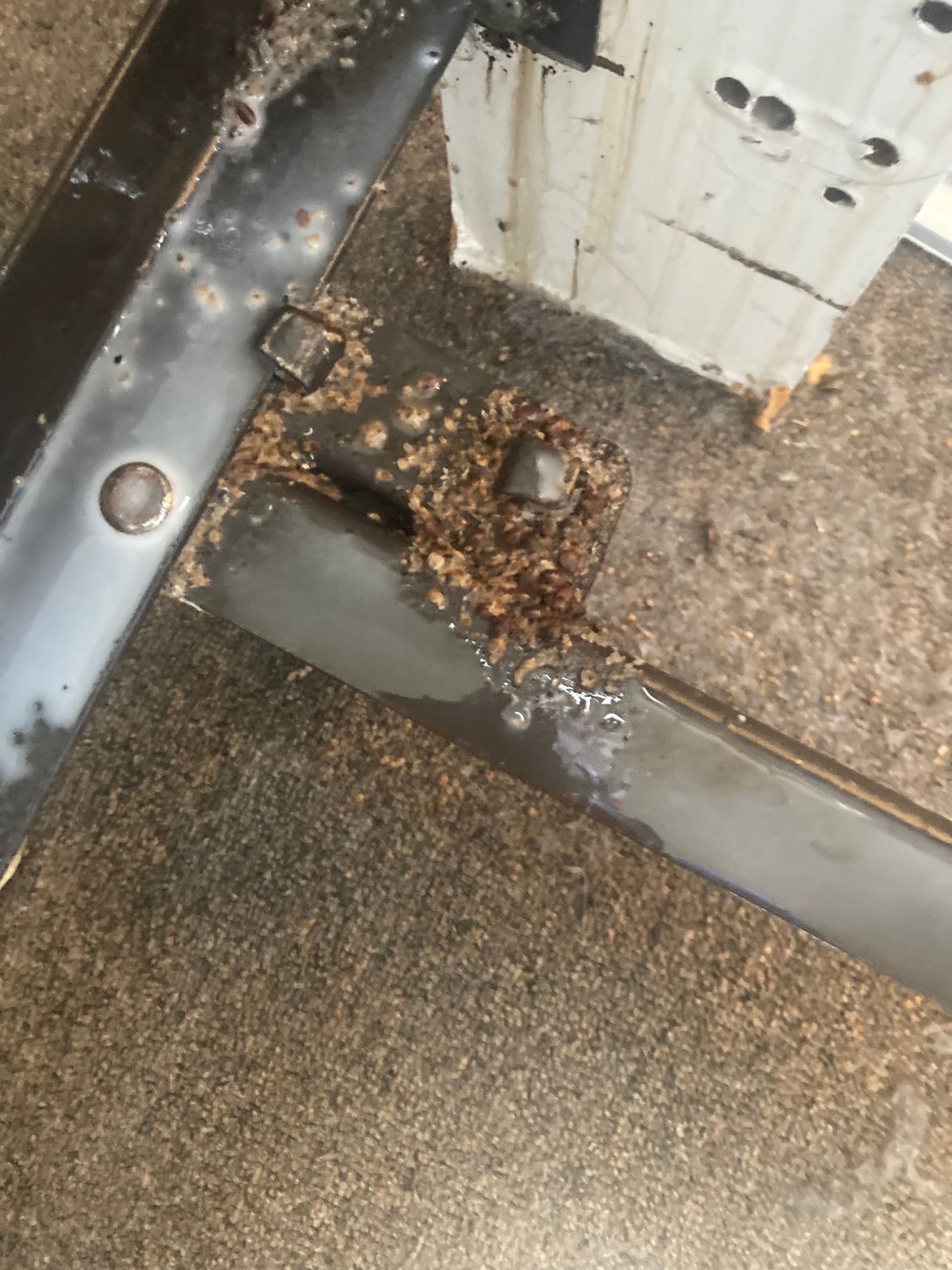 Close-up of rusted metal frame corner with welding and debris, on a brown surface, near a white wall.