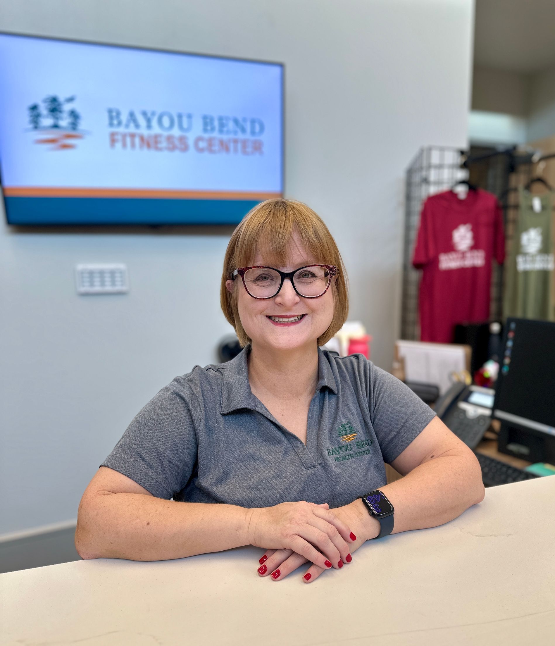 A woman wearing glasses is sitting at a desk in front of a bayou bend fitness center.