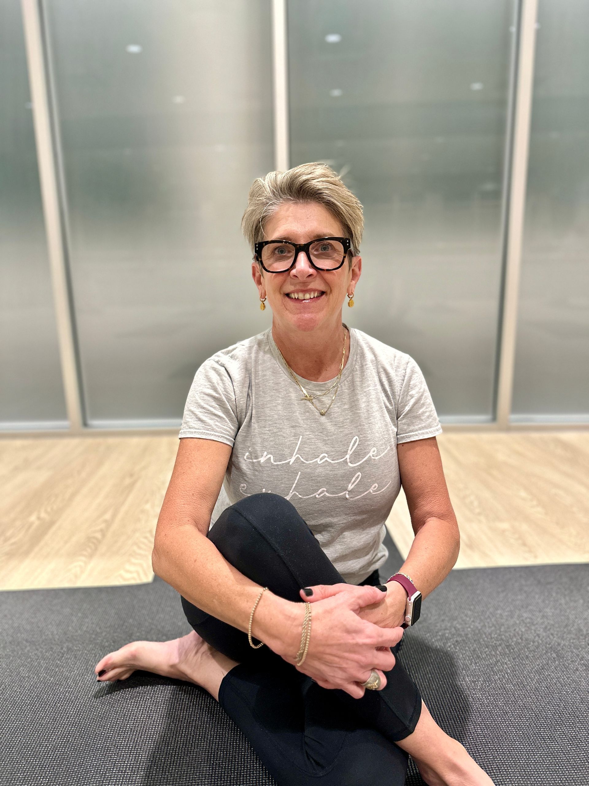 A woman is sitting on a yoga mat with her legs crossed and smiling.