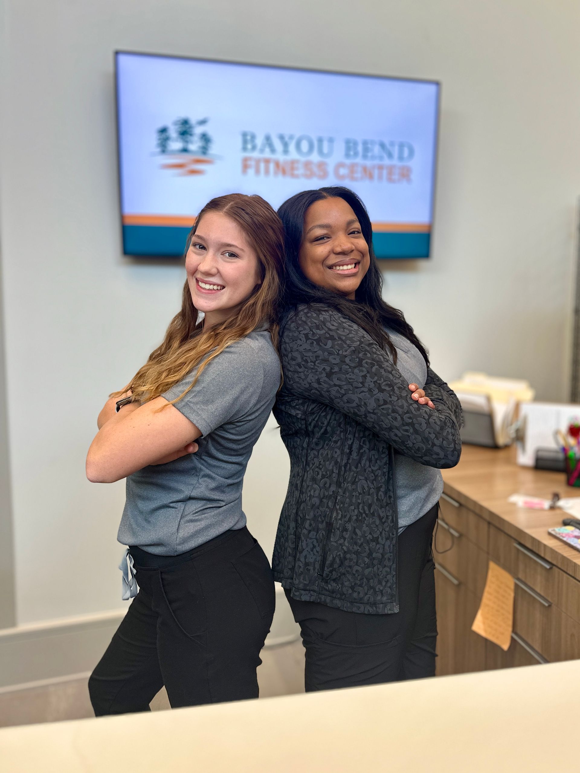 Two women are posing for a picture in front of a bayou bend fitness center sign.