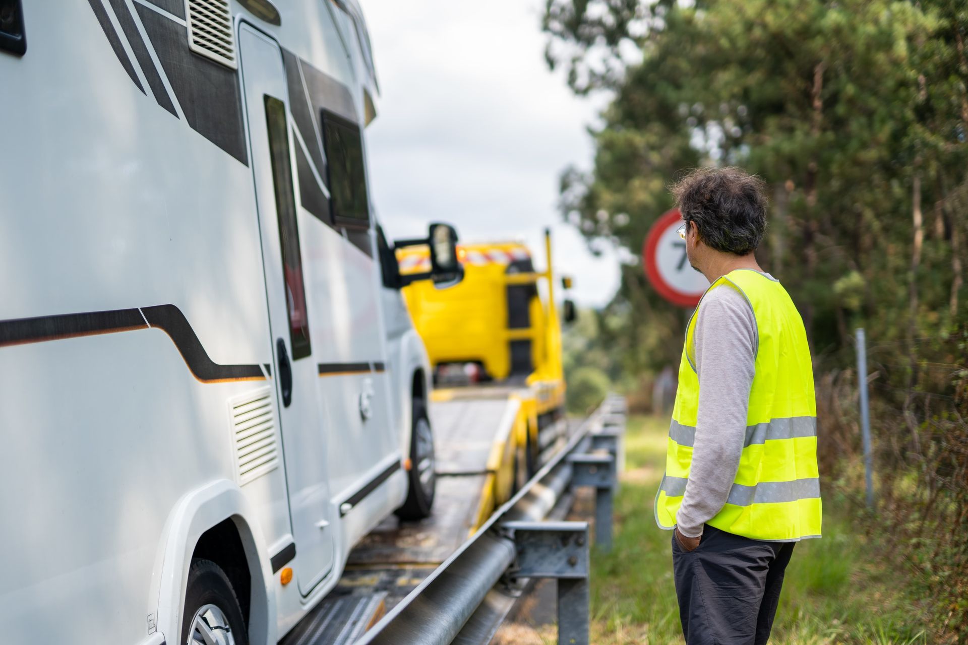Man in safety vest watches RV being towed by a yellow tow truck on a roadside.