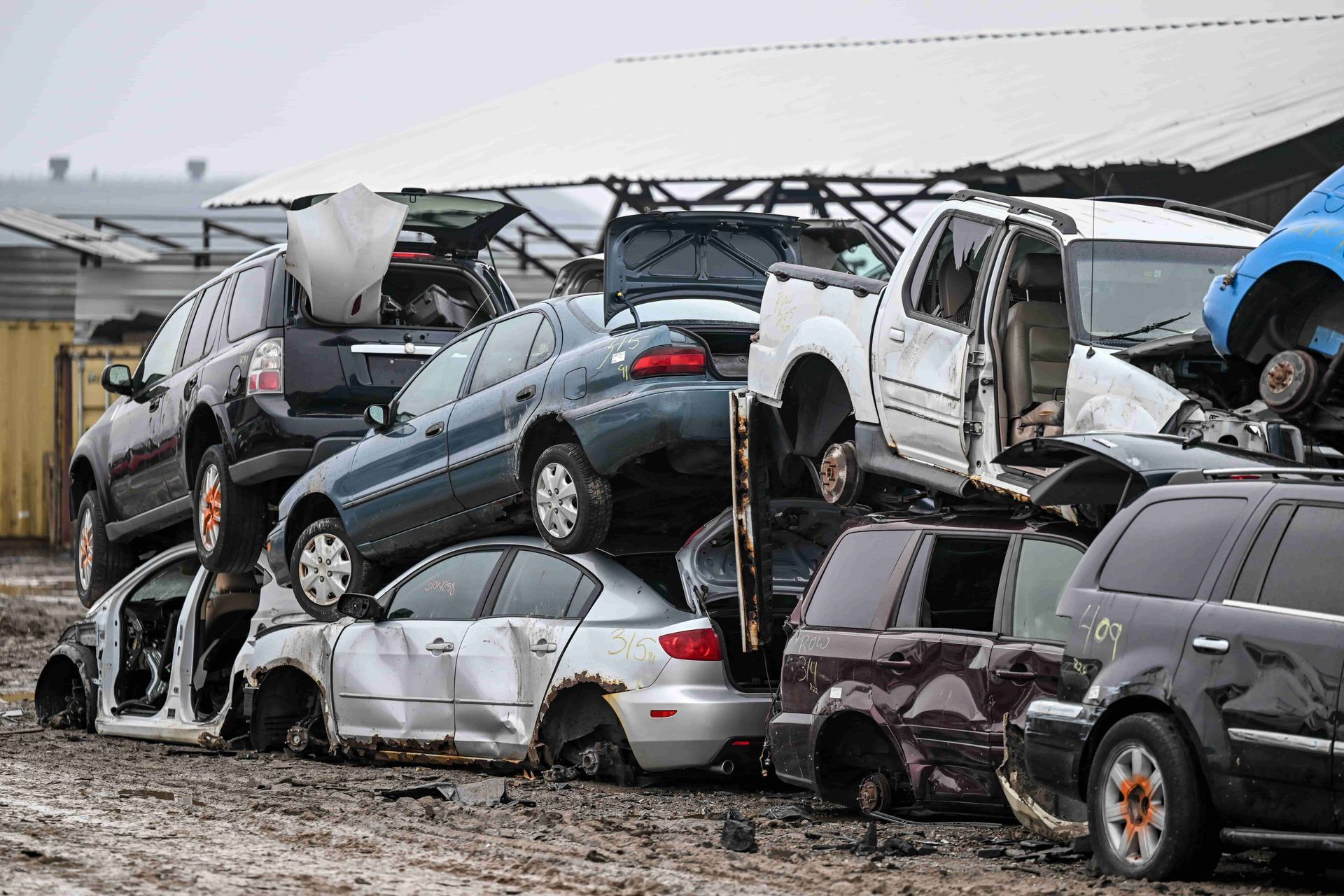 Pile of crushed cars in a junkyard; vehicles are stacked high in muddy lot.