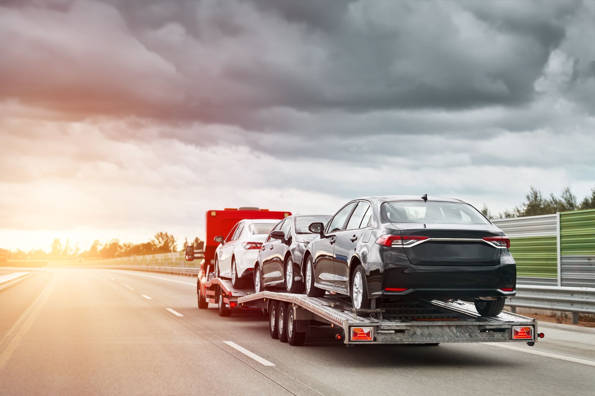 Car carrier truck transporting several cars on a highway with a cloudy sky and sunset.