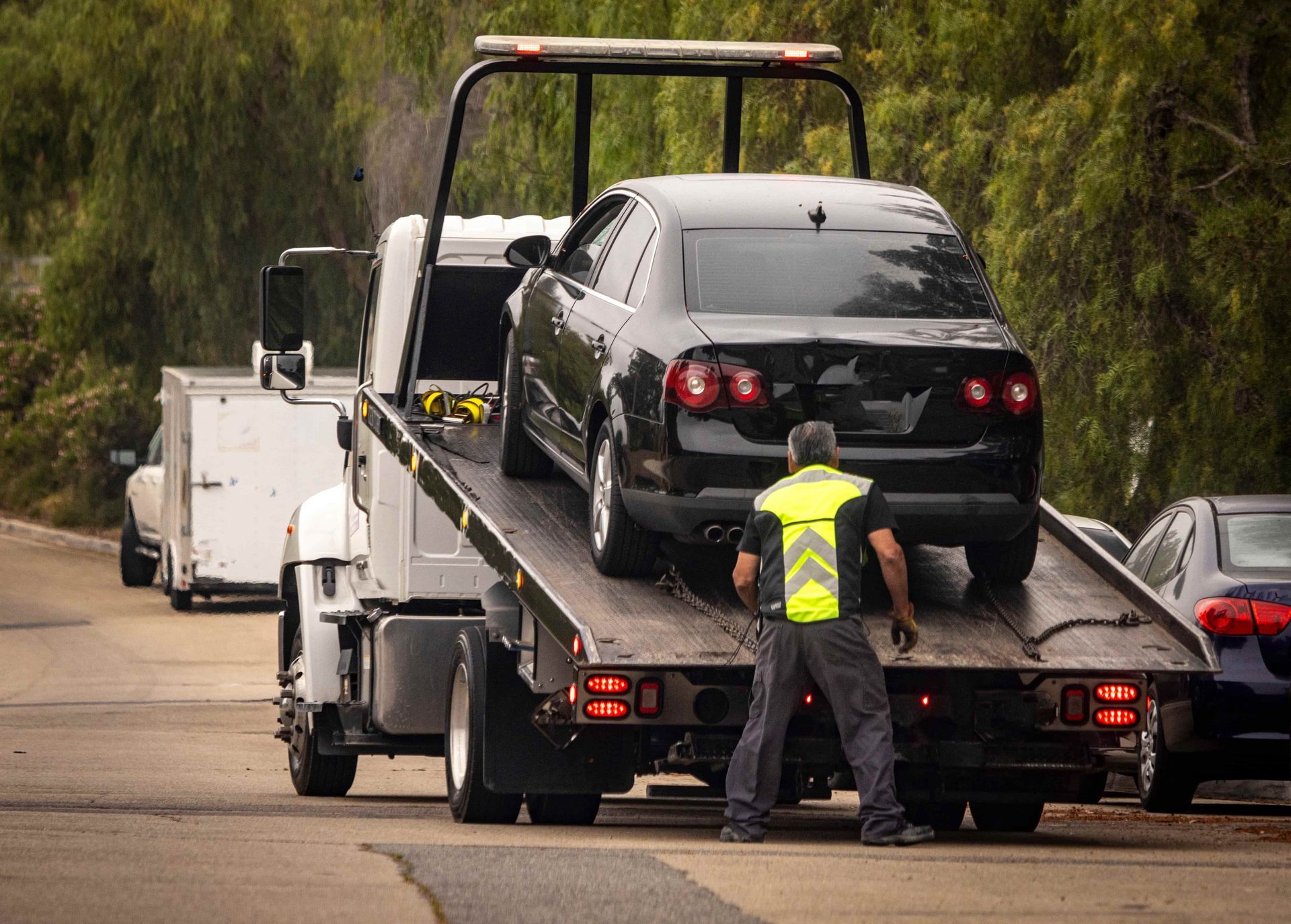 Black car being towed by a tow truck on a road; a worker in a safety vest guides the process.