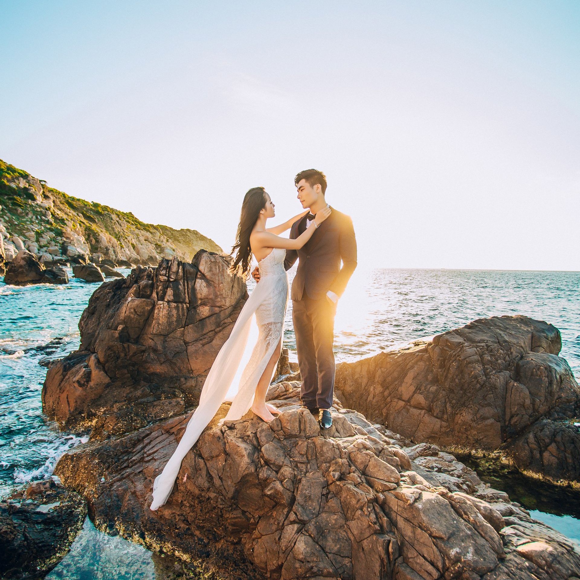 a bride and groom are standing on a rock near the ocean .