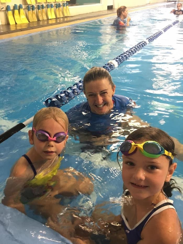 A Swim Instructor Smiles in a Pool Beside Two Young Children Wearing Colorful Goggles