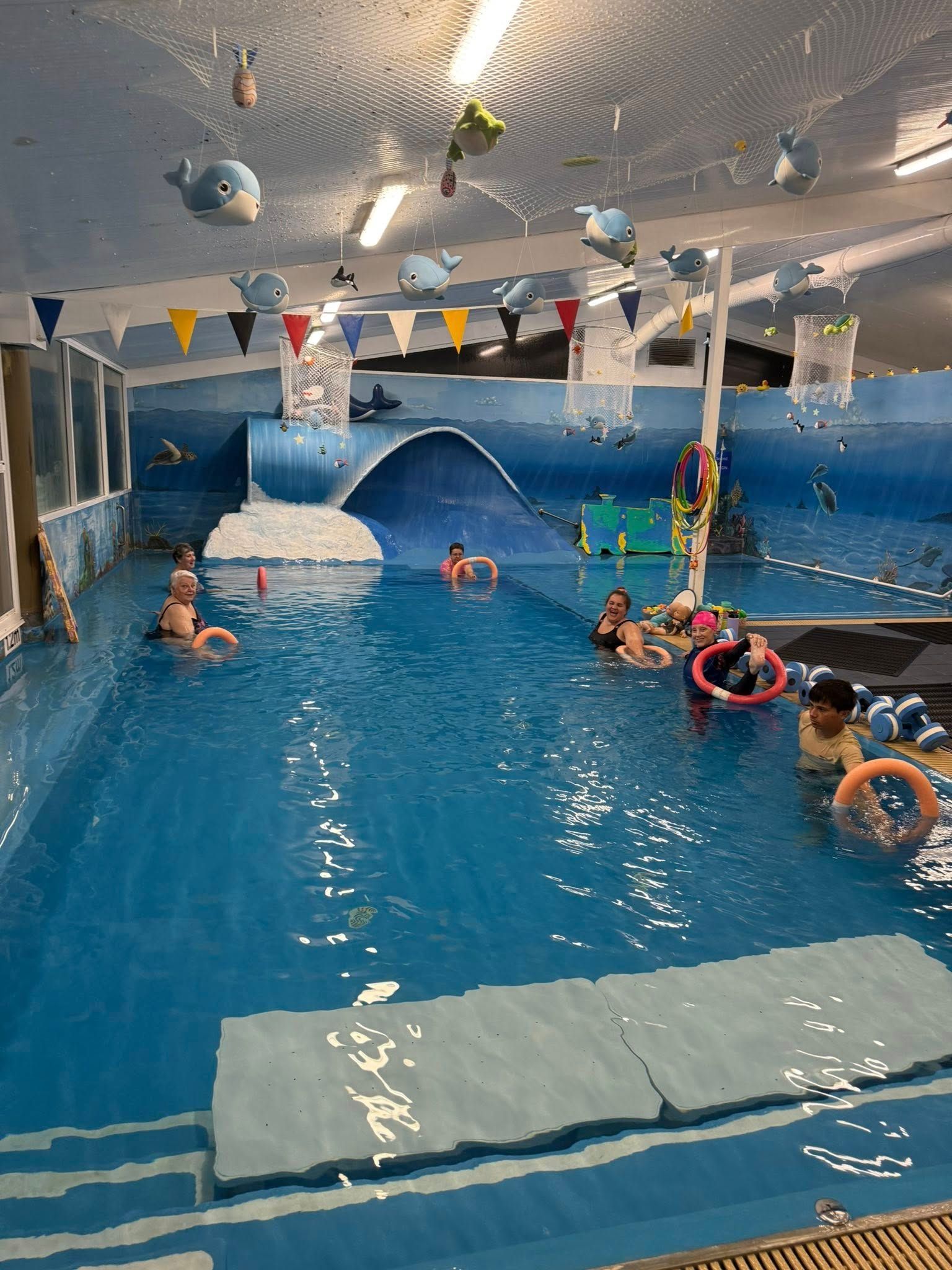 Children Swim in a Blue Indoor Pool, Some With Floats