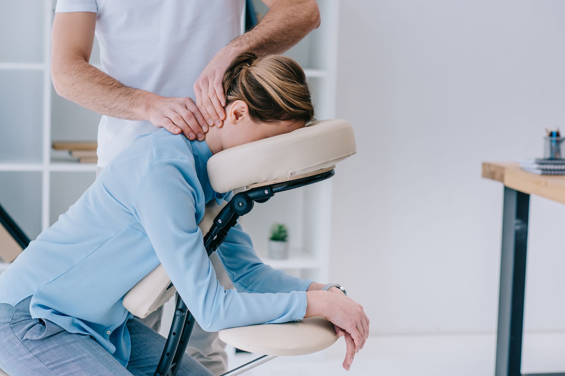 A massage therapist massages the neck of a person resting their head in a padded support on a massage chair.