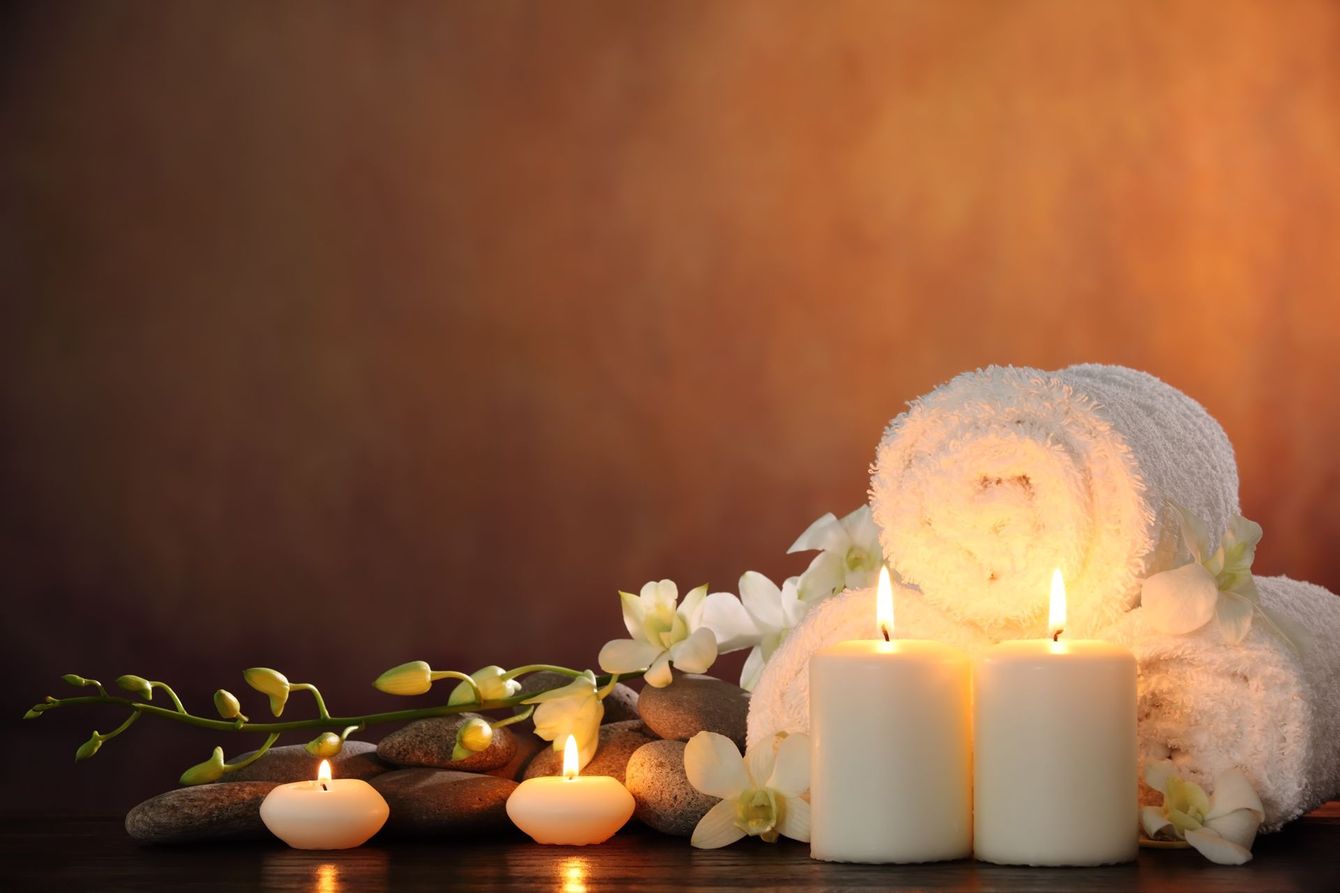 Two white candles, rolled towels, and a delicate flower stem resting on smooth stones against an orange-brown backdrop.