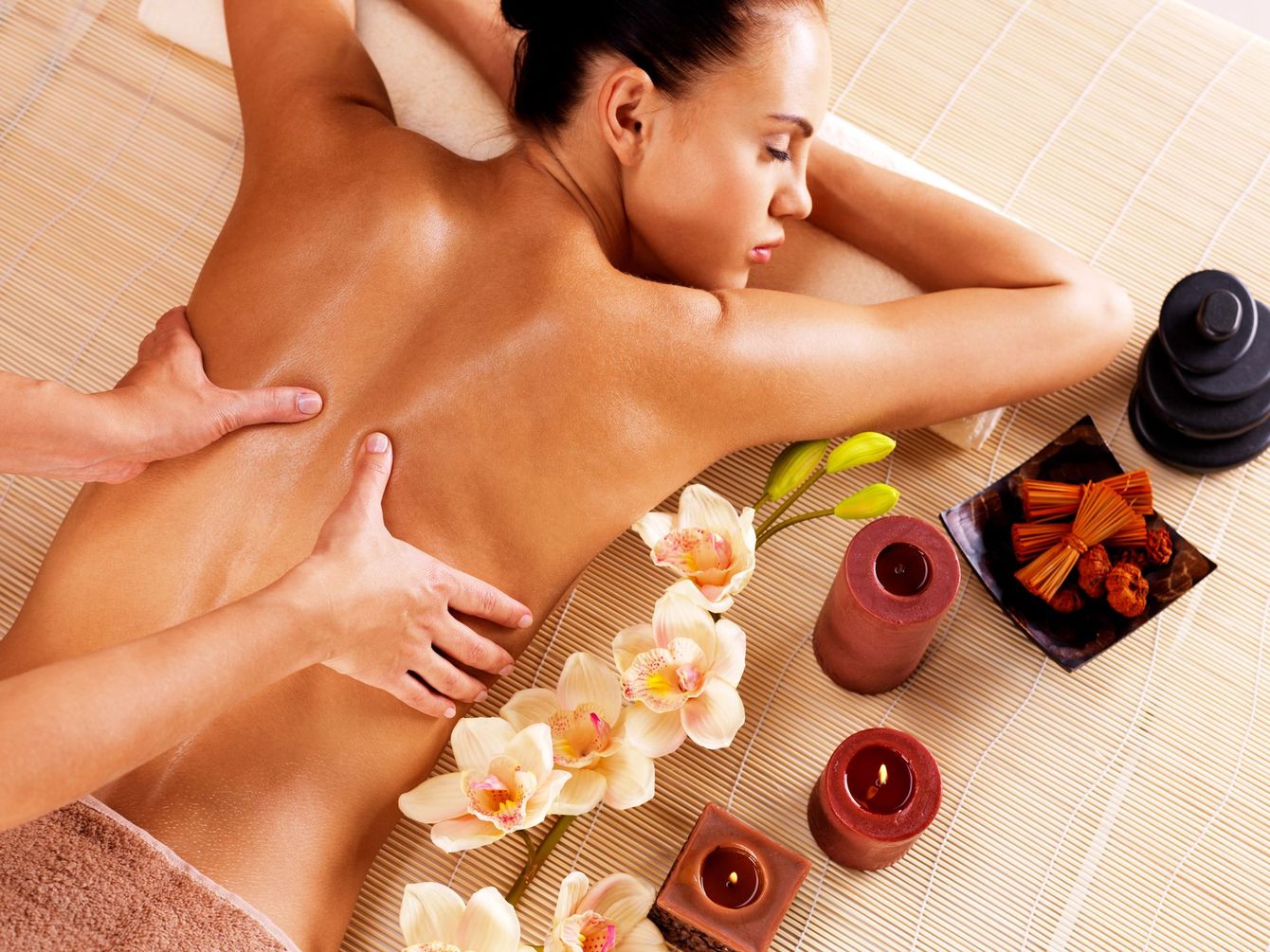 A person receives a back massage in a spa, with orchids, candles, and stacked stones on a bamboo mat nearby.