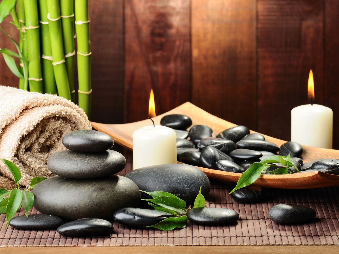 A spa setting with stacked black stones, bamboo stalks, a rolled towel, and two lit candles on a wooden mat.