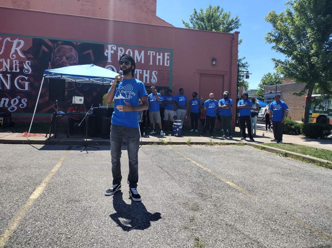 Man speaks into a microphone at an outdoor event, a group of people in blue shirts stand in background.