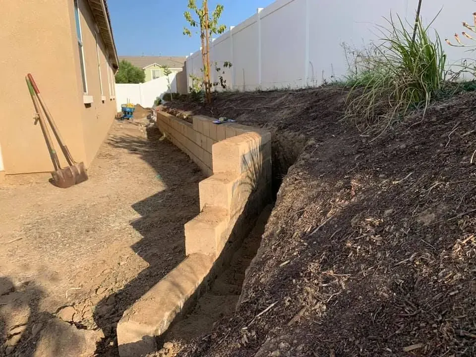 A concrete wall is being built in a backyard next to a white fence.