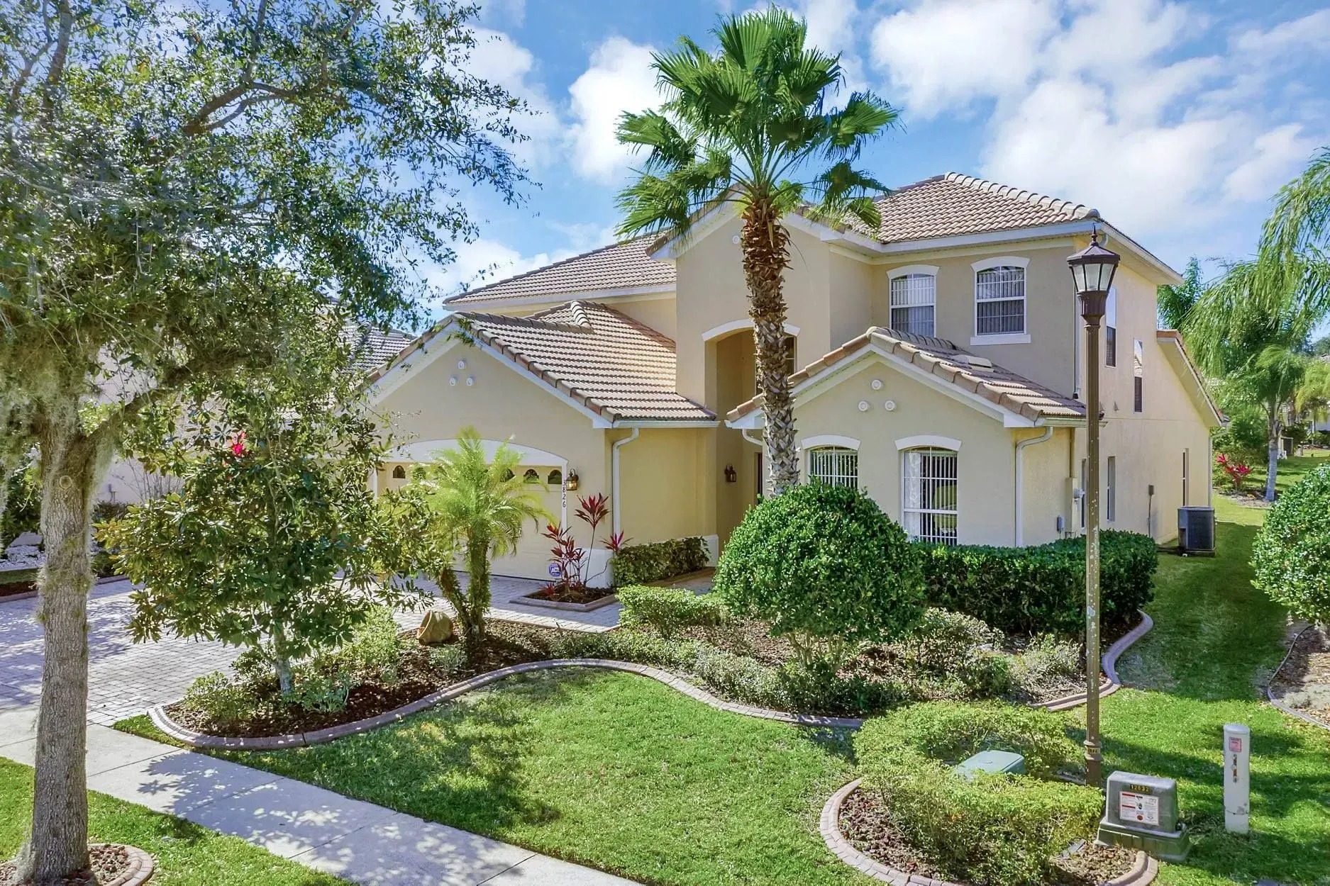 A beige, two-story suburban home with a tile roof, palm tree, and landscaped front yard under a blue sky.