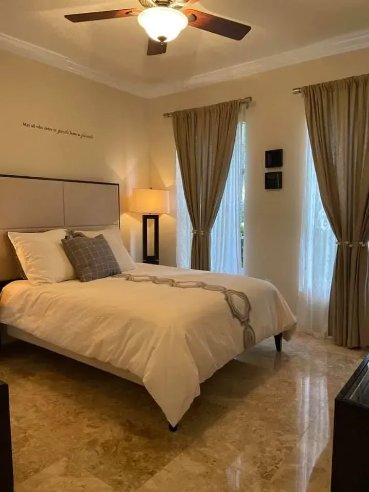 A bright bedroom with a white-covered bed, two large windows with tan curtains, a ceiling fan, and tiled flooring.