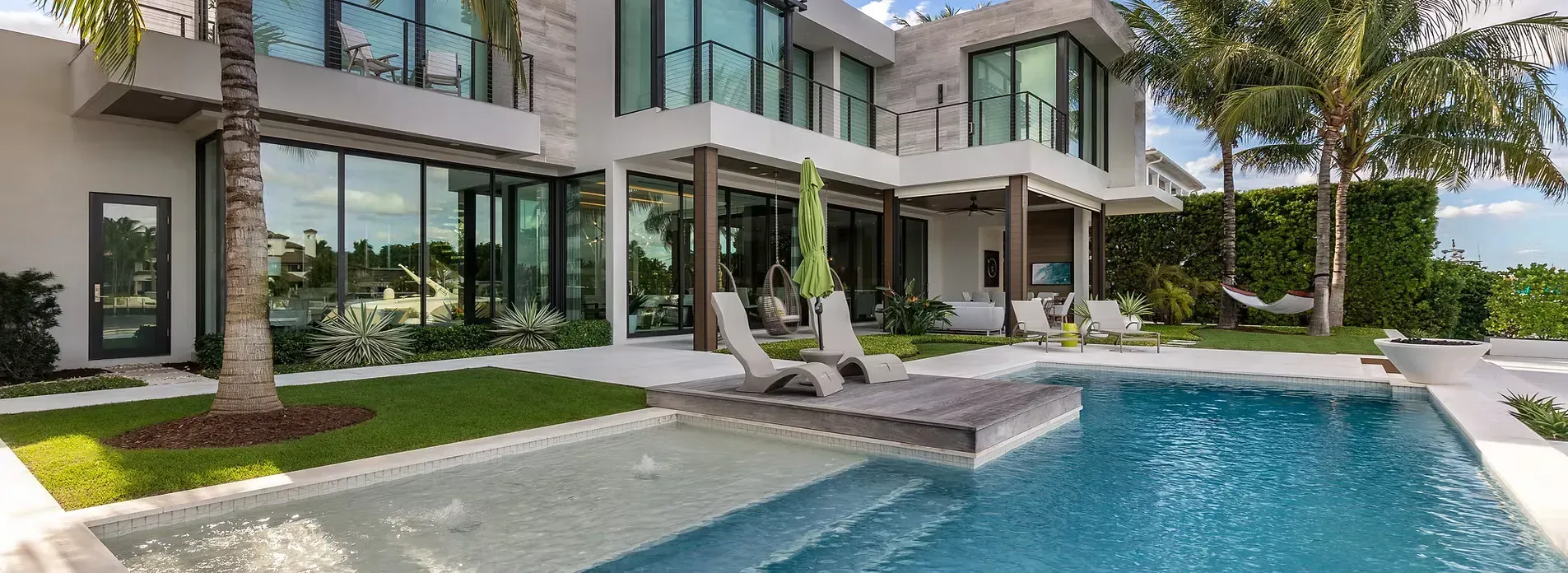A modern luxury house with large glass windows, a swimming pool, and lounge chairs on a patio with palm trees.