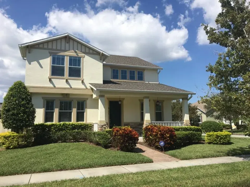 A two-story, cream-colored suburban house with a front porch, tan trim, and landscaped bushes under a blue, cloudy sky.