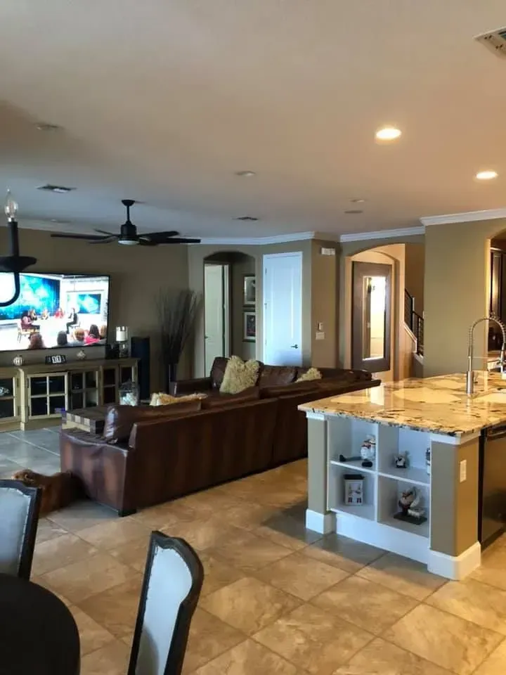 An open-plan living room and kitchen featuring a brown sectional sofa, a large television, and a granite-topped island.
