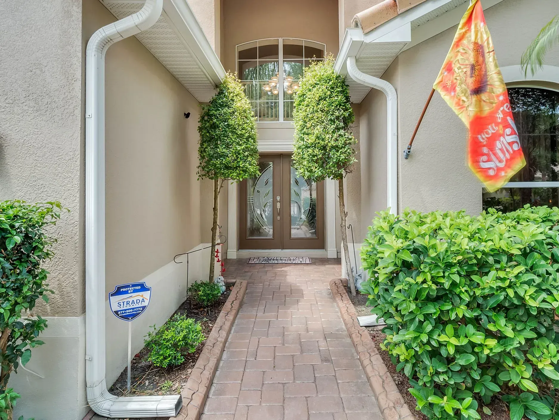 A brick walkway leads to double front doors, framed by two tall, manicured shrubs and a small orange flag on the wall.