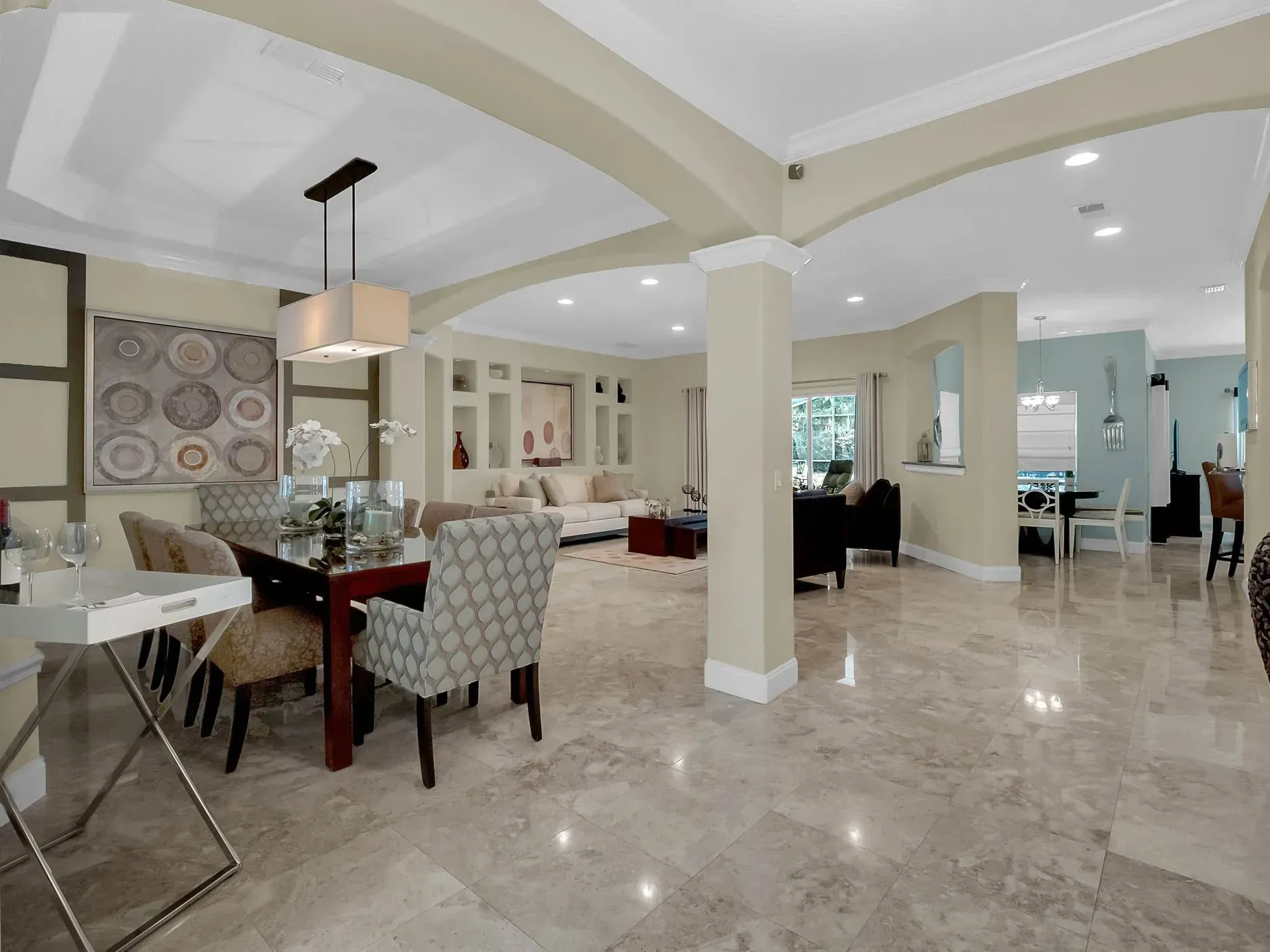 A brightly lit, open-concept dining room with marble floors, a patterned chair set, and a living area in the background.