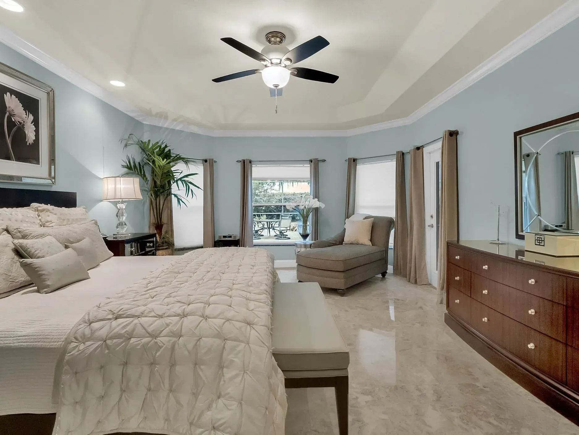 A spacious bedroom with light blue walls, marble floors, a king bed, a gray chaise lounge, and a dark wood dresser.