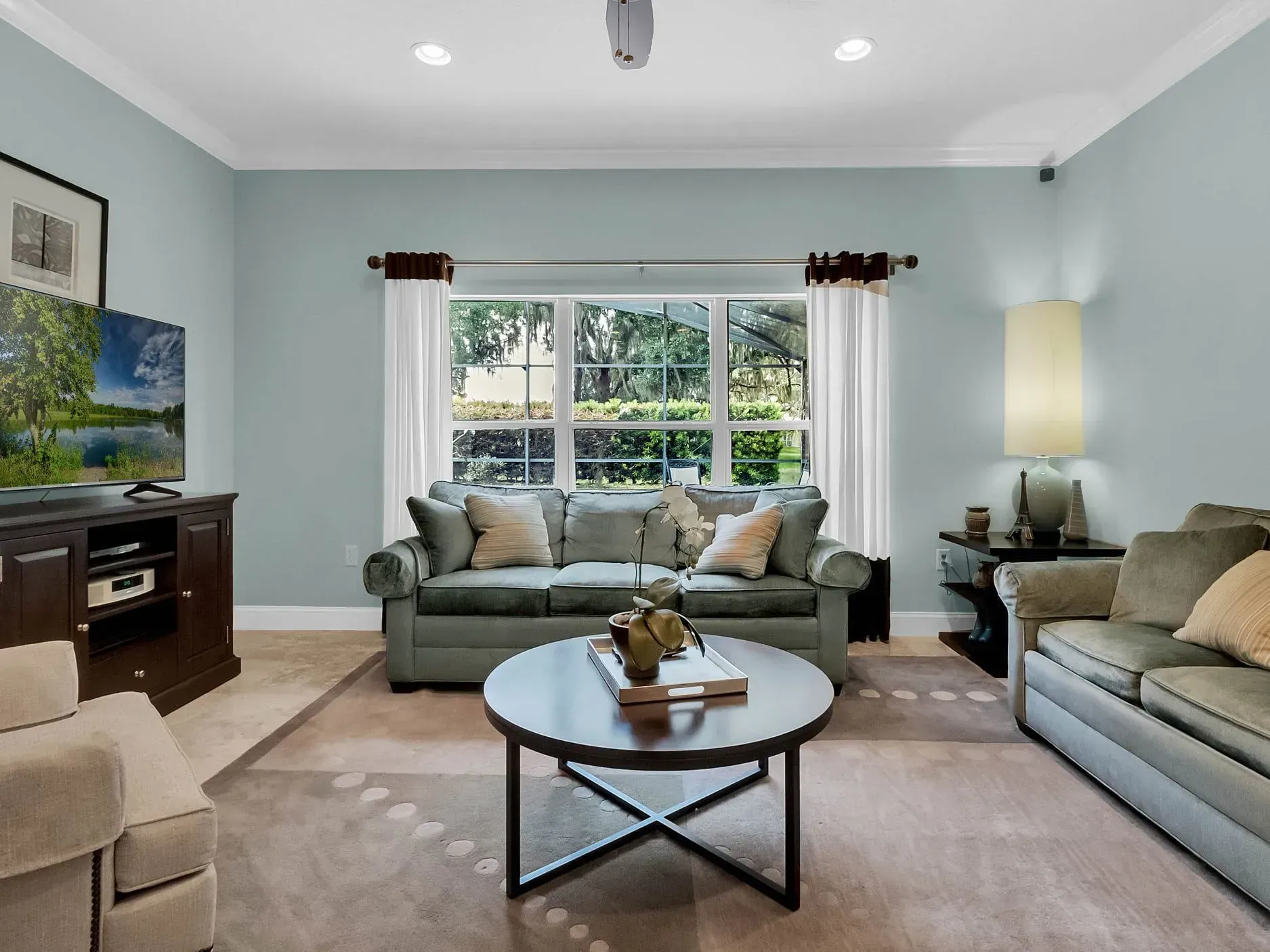 A living room with pale blue walls, a sofa facing a window, a round coffee table, and a TV on a dark wooden stand.
