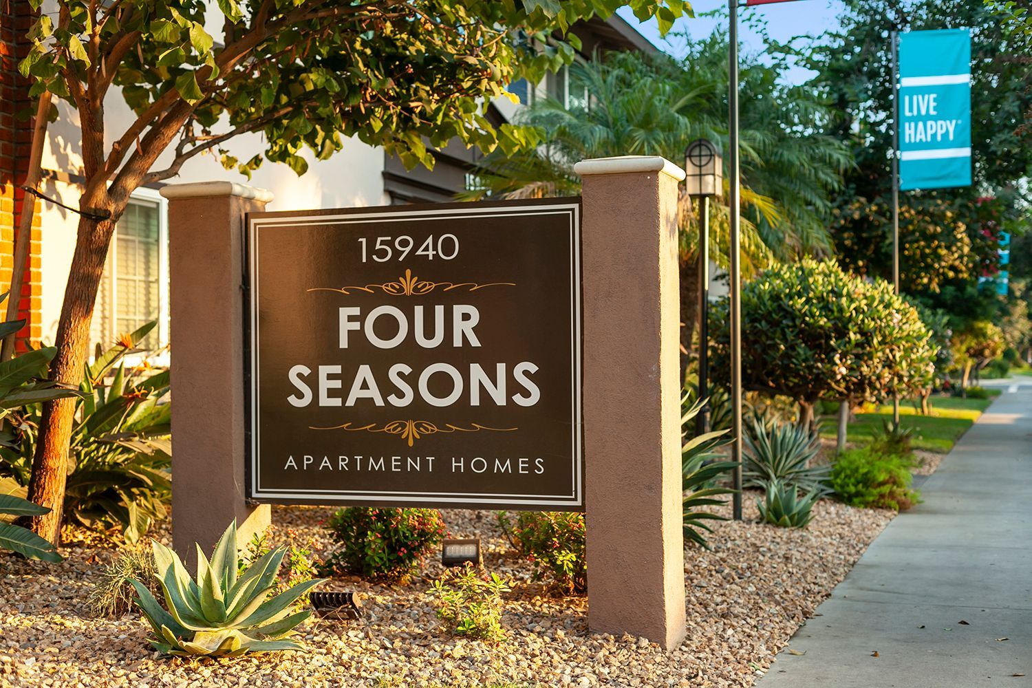 Monument sign of Four Seasons Apartment Homes with a blue live happy flag and landscape in background