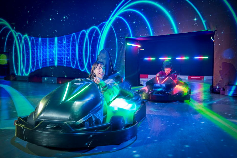 Two people race electric go-karts on a track with neon blue and green lights.