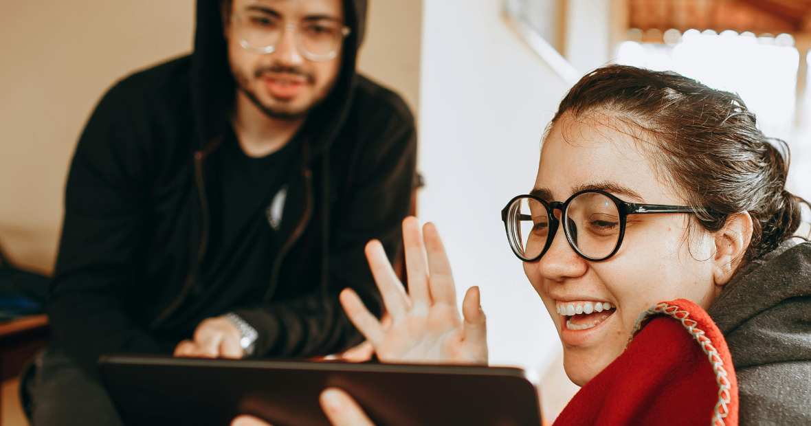 Woman with glasses waves while looking at a tablet, smiling. Man in a hoodie looks on.