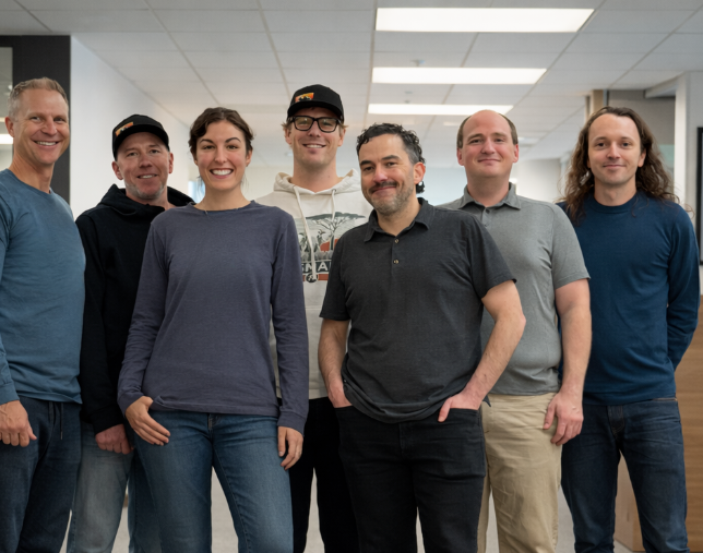 Group of people in casual attire standing in an office, smiling at the camera.