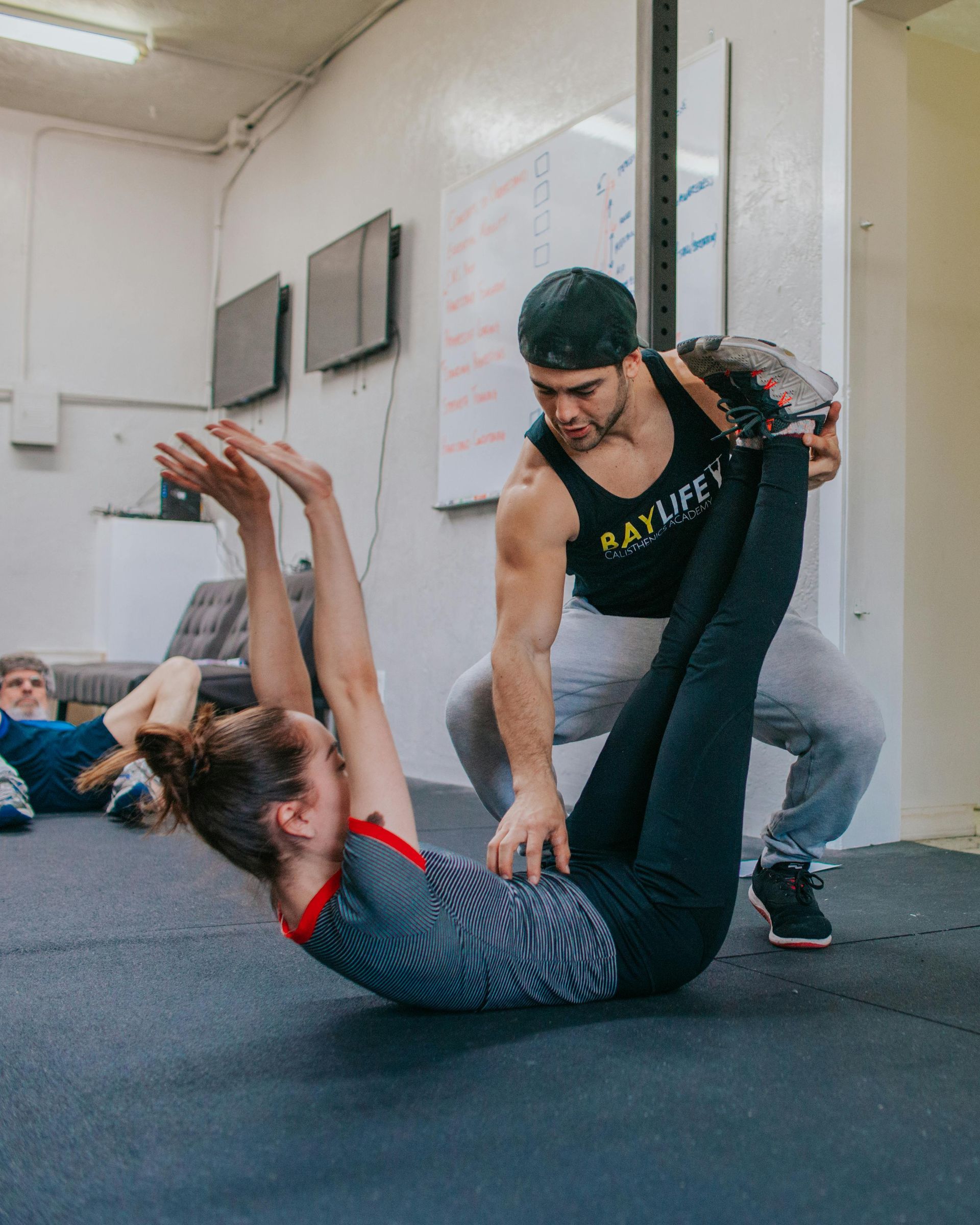 A fitness instructor assists a woman performing a leg-raise exercise on a gym floor. The woman is in a reclined position.