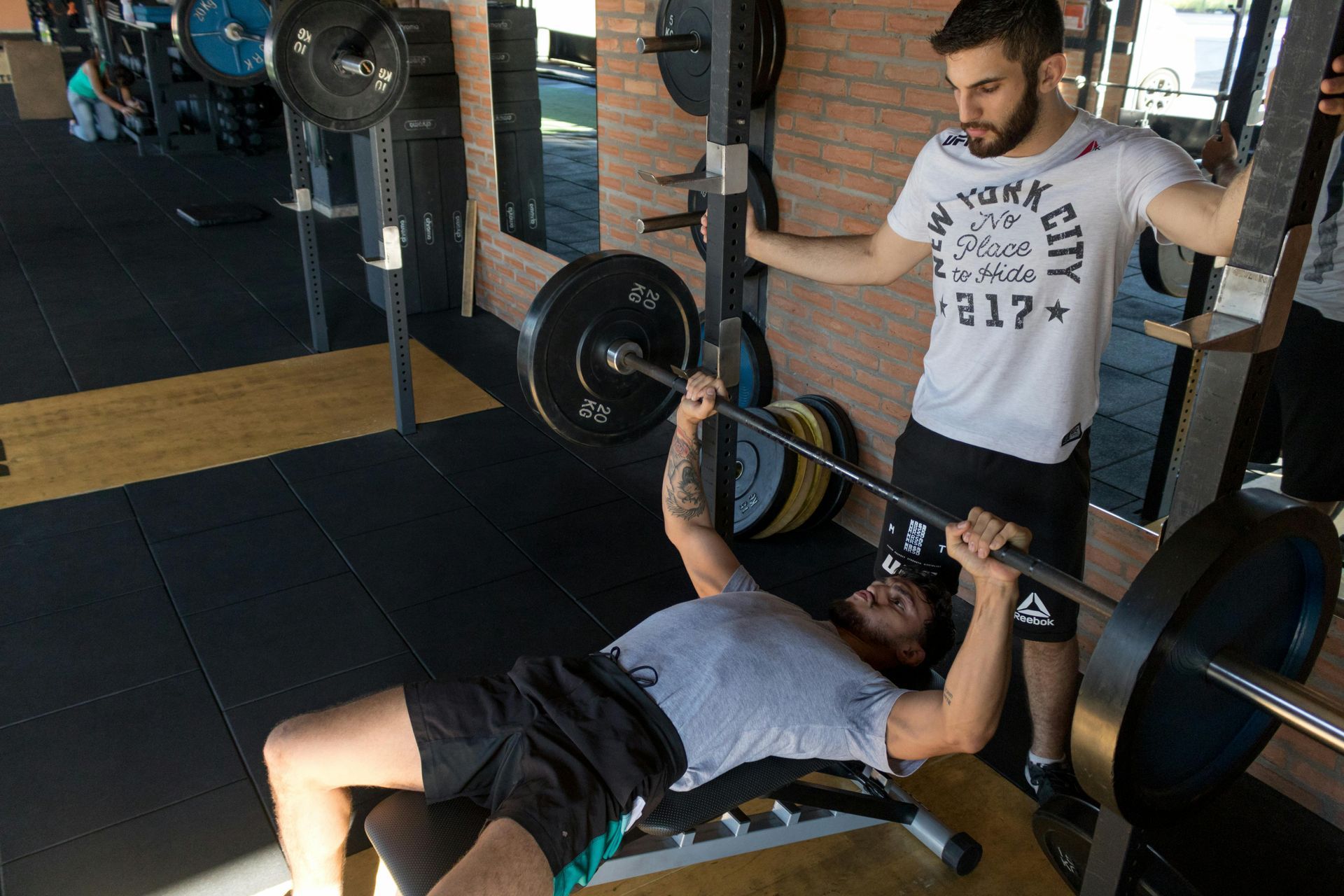 Man bench presses with spotter at a gym. The lifter is wearing a grey shirt and black shorts, the spotter is in a white shirt and black shorts.