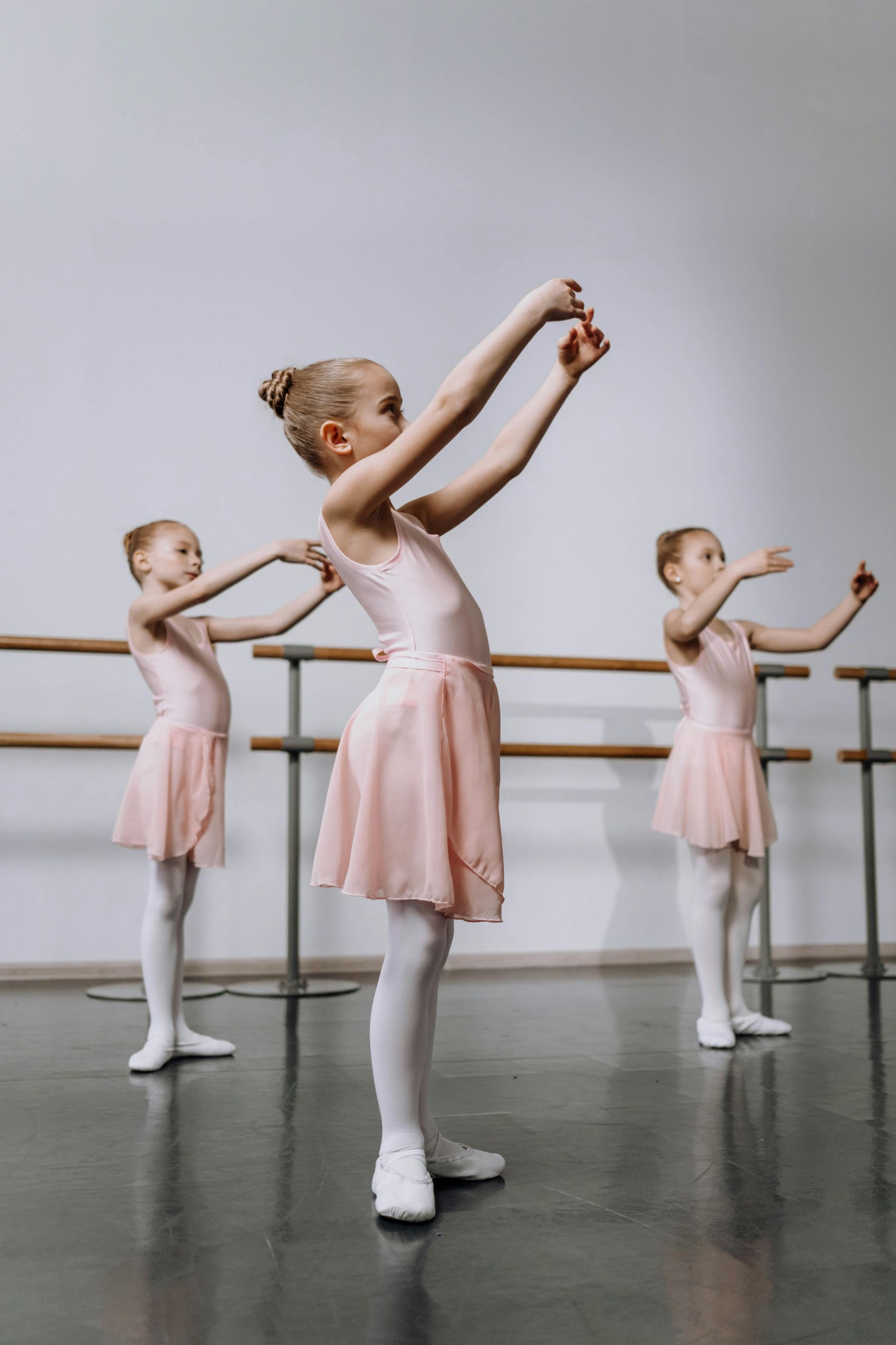 Three young ballerinas in pink leotards and tutus practice at the barre in a dance studio.