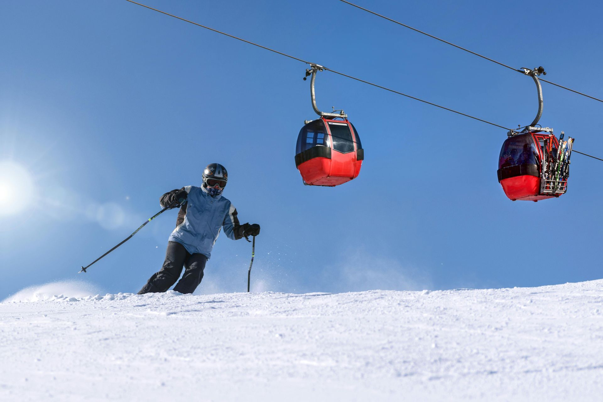 Skier on snowy slope, descending past red cable car; sunny, blue sky.