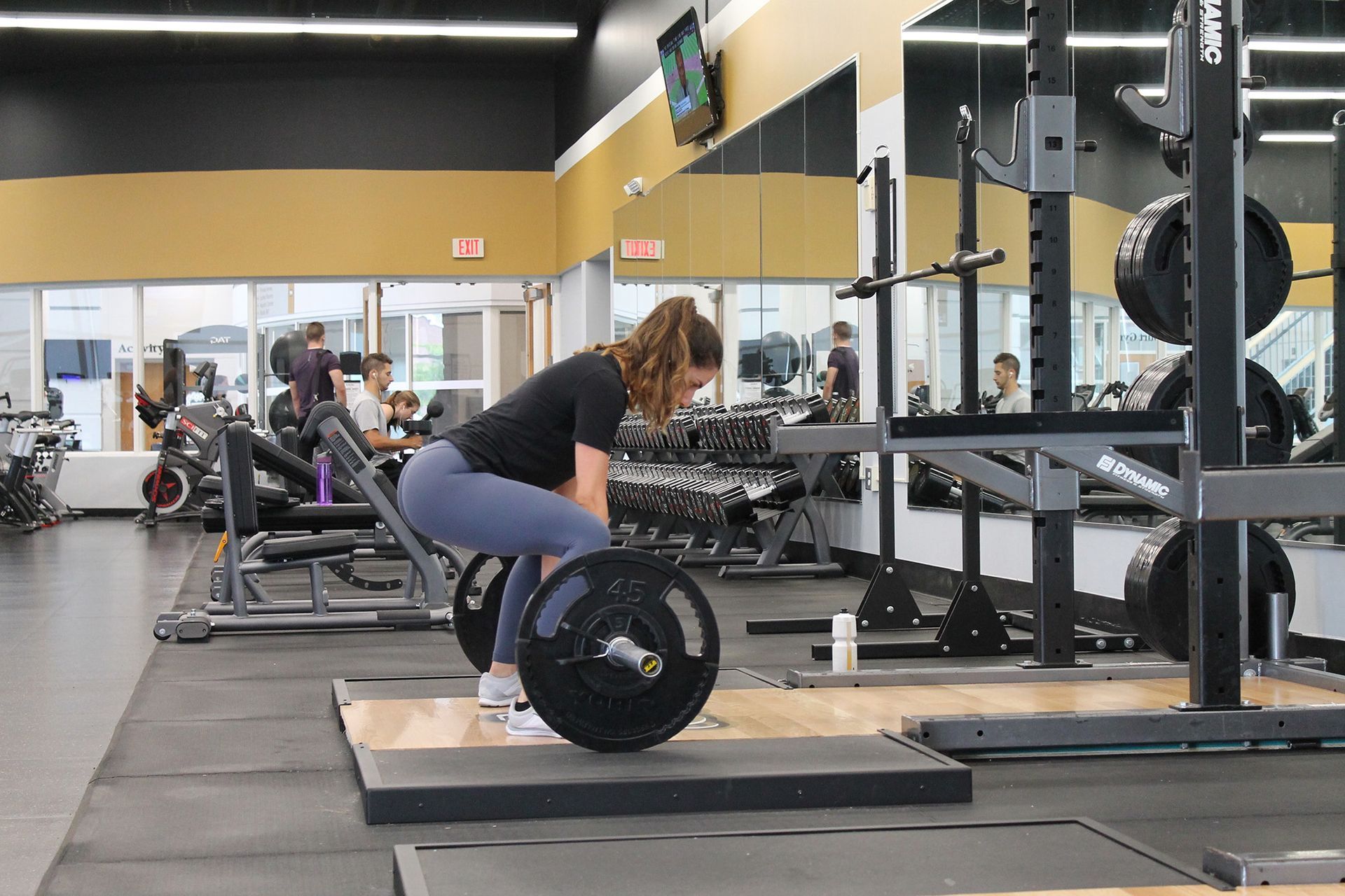 Woman performing a deadlift in a gym, barbell on floor. She's bent over with a neutral spine, wearing black shirt, blue leggings, and white shoes.
