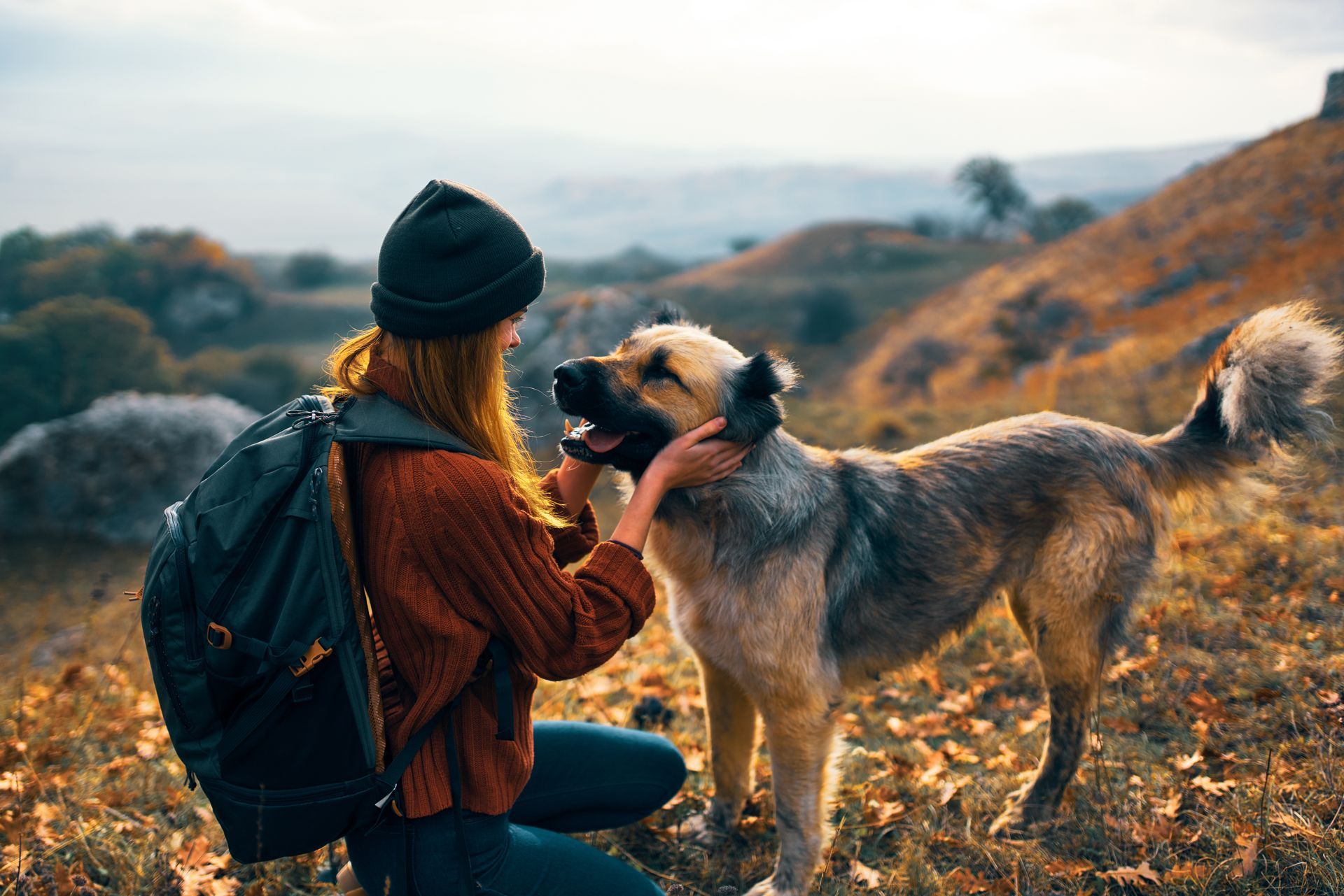 A woman with a backpack is petting a dog on a hill.
