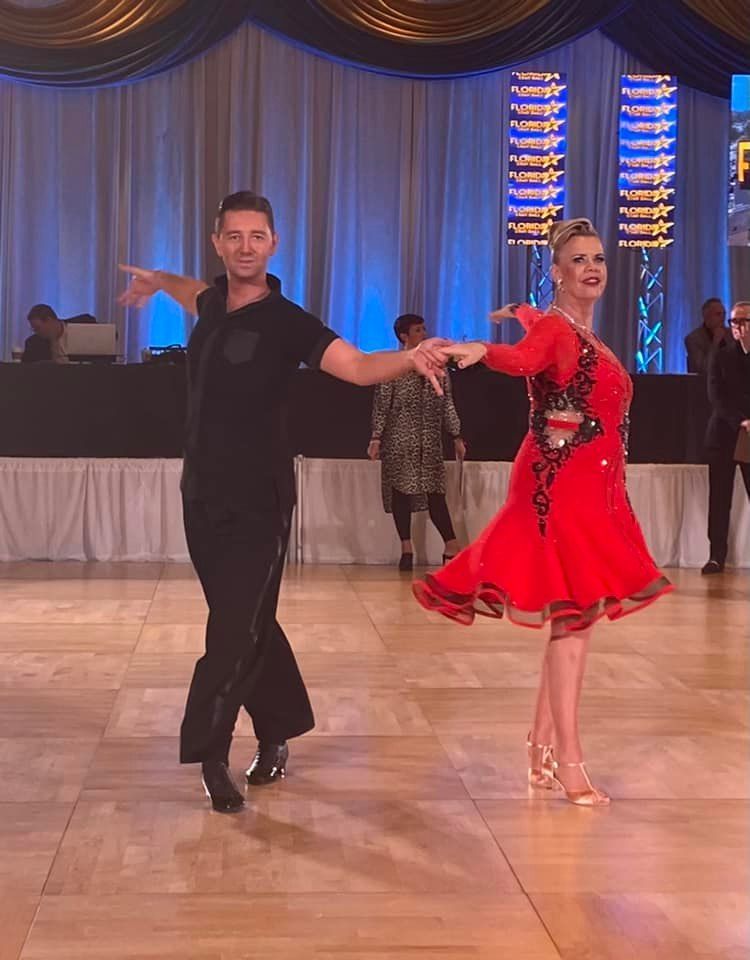 A man and woman ballroom dancing on a wooden floor, she in a red dress, he in black, at a competition.