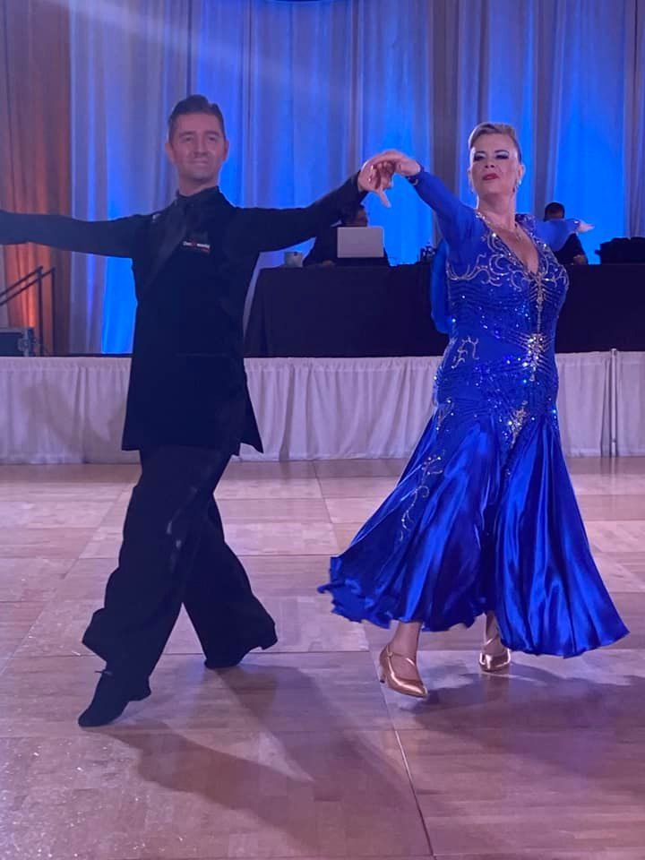 A man and woman ballroom dance. The woman wears a blue dress; the man, a black suit. They perform on a stage.