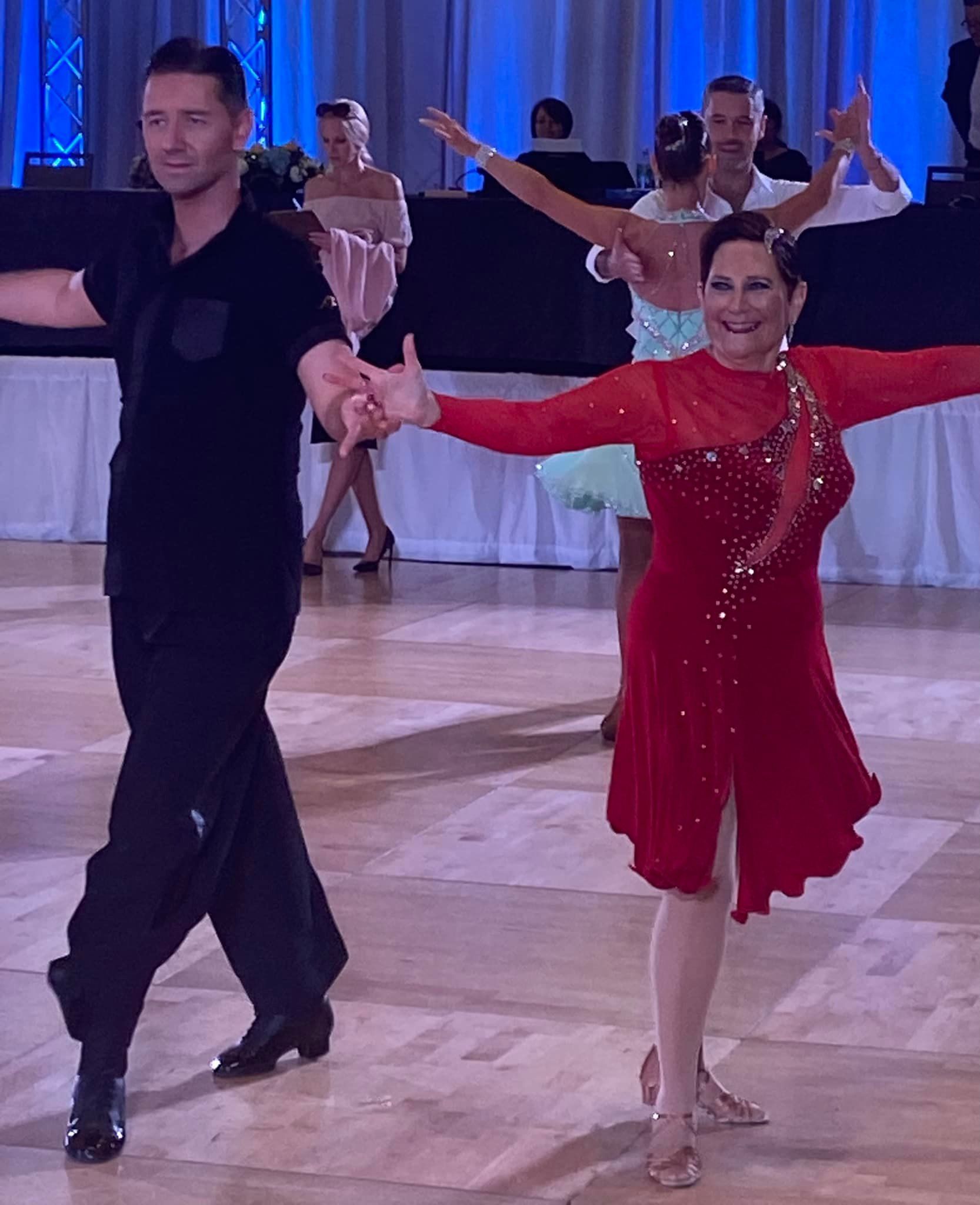 Couple dancing in a ballroom, man in black, woman in red dress. Other dancers in the background.